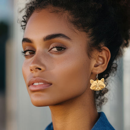 A close-up portrait of a woman wearing a gold fox-shaped dangle earring, set against a softly blurred neutral background.