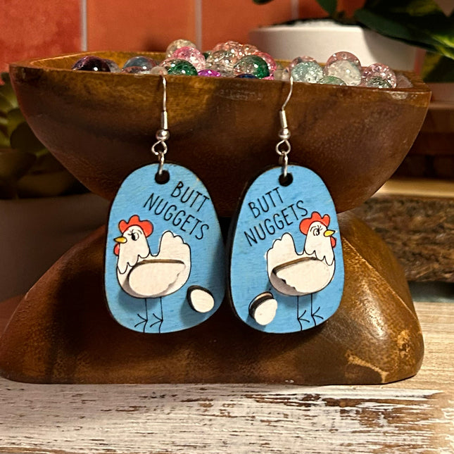 Close-up of hand-painted birchwood "Butt Nuggets" chicken earrings displayed on a wooden stand