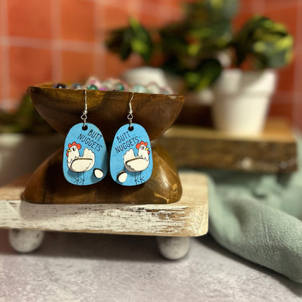 Hand-painted birchwood "Butt Nuggets" chicken earrings with sterling silver fishhook wires, displayed on a tiered wood stand