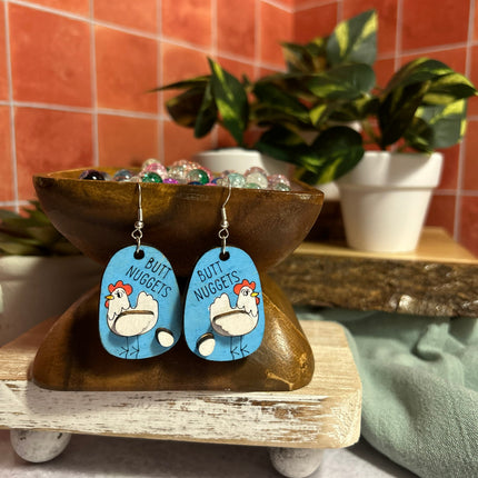 Hand-painted birchwood "Butt Nuggets" chicken earrings displayed on a wooden stand with colorful glass beads and potted plants in background