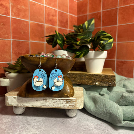 Hand-painted birchwood "Butt Nuggets" chicken earrings on a tiered wood stand with a terracotta tile wall in background