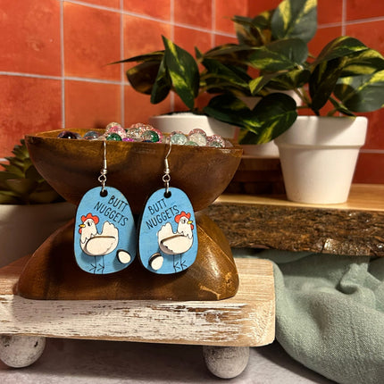 Hand-painted birchwood "Butt Nuggets" chicken earrings displayed on a wooden stand with beads and potted plants