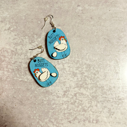 Pair of hand-painted birchwood "Butt Nuggets" chicken earrings laid flat on a stone surface