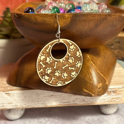 A single round wooden earring with paw print and bone engravings, hanging from a silver fishhook wire, displayed in front of a wooden bowl of colorful beads.