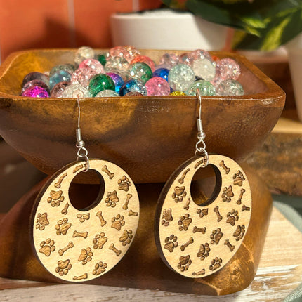 A pair of oval wooden drop earrings with paw print and bone patterns, displayed in front of a wooden bowl filled with colorful glass beads.