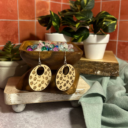 A pair of oval drop earrings with beige floral-patterned wooden discs and silver fishhook wires, displayed on a rustic wooden stand with decorative plants and a terracotta tiled background.