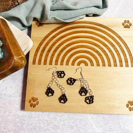 A pair of dangling earrings displayed on a wooden stand. The earrings have black paw-print charms and silver-tone chain links, resting on a light wood board with engraved paw prints and a rainbow design.