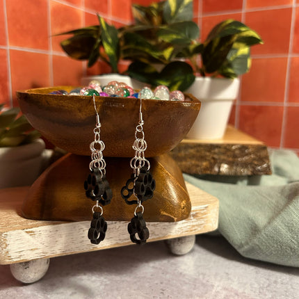 A pair of handmade dangle earrings displayed on a rustic wooden stand. The earrings feature silver-toned metal rings and black flower-shaped charms, set against a styled background with a terracotta tiled wall, potted plants, and a sage green cloth.