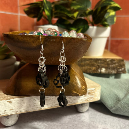 A pair of handmade dangle earrings with silver chain links and black paw-shaped charms, displayed on a rustic wooden stand with decorative plants in the background.