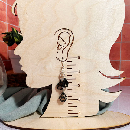 A single dangling earring displayed on a wooden head-shaped stand with an ear measurement scale. The earring has a silver-toned metal top, a clear faceted bead, a black bead, and a black paw-print charm.
