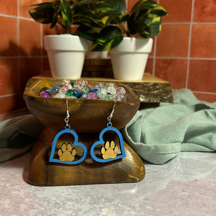 Blue hand-painted birchwood paw heart earrings displayed on wooden bowl stand with potted plants in background