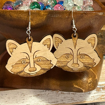 A pair of wooden raccoon face dangle earrings with laser-engraved details, hanging from silver fishhook wires, displayed in front of a rustic wooden bowl filled with colorful glass beads.