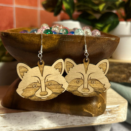 A pair of wooden raccoon face dangle earrings displayed on a rustic wooden stand. The earrings are made from light natural wood with darker brown laser-engraved facial details and hang from silver-toned fishhook wires.