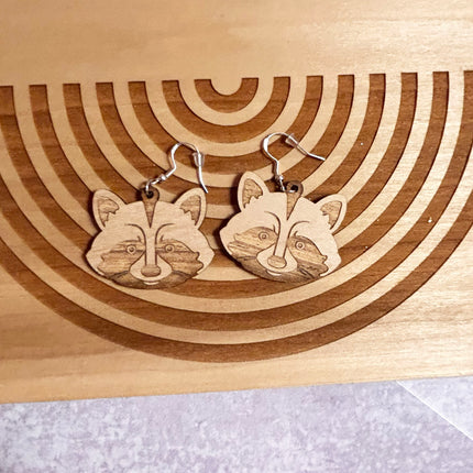 A pair of wooden raccoon face dangle earrings with silver fishhook wires, displayed on a wooden board with concentric circular laser-cut grooves.