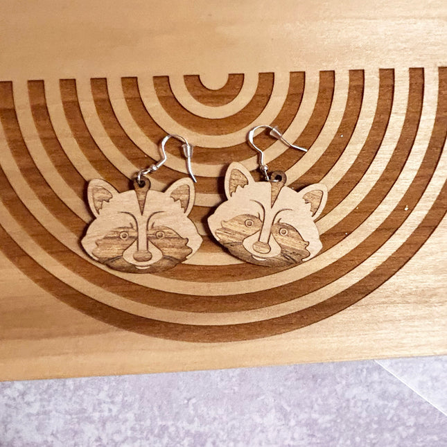 A pair of wooden raccoon face dangle earrings with silver fishhook wires, displayed on a wooden board with concentric circular laser-cut grooves.