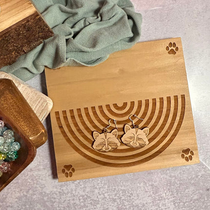 A pair of wooden cat-shaped dangle earrings displayed on a laser-engraved wooden stand, styled with a sage green cloth and a small wooden tray on a gray textured surface.
