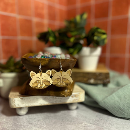 A pair of wooden raccoon face dangle earrings displayed on a rustic wooden stand, with a blurred background of potted plants and terracotta tiles.