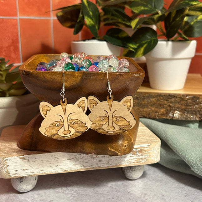 A pair of wooden raccoon face dangle earrings displayed on a decorative wooden stand, with a bowl of colorful beads and potted plants in the background.