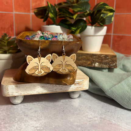 A pair of wooden raccoon face dangle earrings displayed on a rustic wooden stand, with a matching wooden bowl of colorful beads behind them and potted plants in the background.