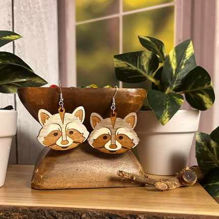 A pair of wooden raccoon face dangle earrings with silver fishhook wires, displayed on a rustic gold-colored scale with potted plants in the background.