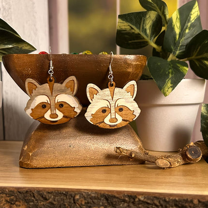 A pair of wooden raccoon-shaped dangle earrings displayed on a rustic wooden stand, with potted green plants in the background.