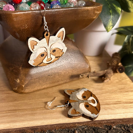 A pair of raccoon-shaped wooden dangle earrings with silver fishhook wires, displayed on a wooden surface with a rustic bowl of colorful beads in the background.