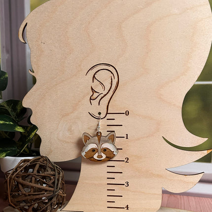 A small raccoon-shaped earring displayed on a wooden ear-measuring prop with millimeter markings. The earring hangs from a silver stud and has tan, cream, and dark brown facial details.