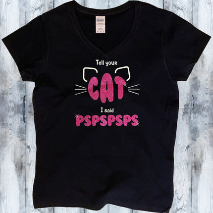 A black short-sleeve V-neck T-shirt laid flat on a light gray wood plank background. The shirt features a graphic print with the text 'Tell your CAT I said PSPSPSPS' in pink and white, accompanied by white cat ear and whisker illustrations.