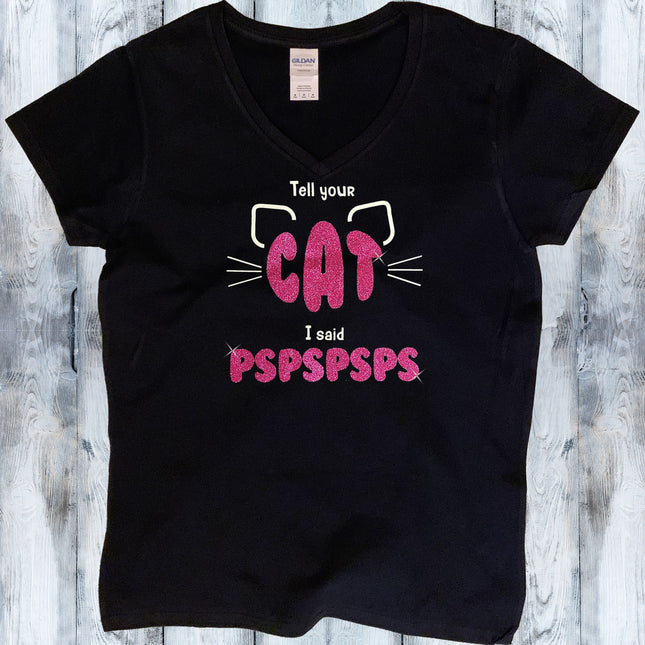 A black short-sleeve V-neck T-shirt laid flat on a light gray wood plank background. The shirt features a graphic print with the text 'Tell your CAT I said PSPSPSPS' in pink and white, accompanied by white cat ear and whisker illustrations.